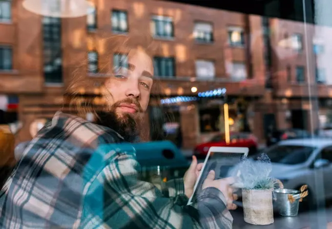 Portrait of stylish young man behind windowpane with tablet in a cafe