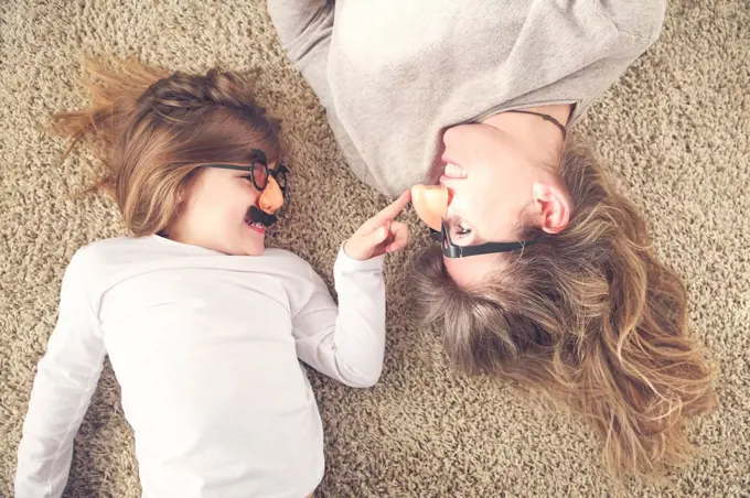 Mother and little daughter lying on the carpet having fun with comedy glasses