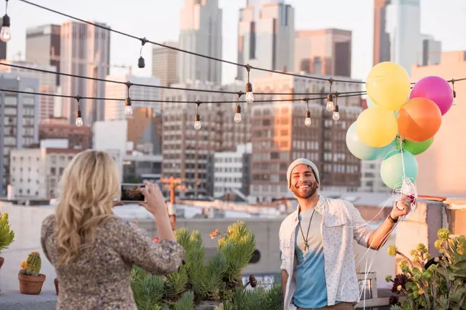 Young couple with balloons standing on rooftop terrace, using smart phone