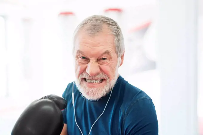 Portrait of aggressive senior man with earphones and boxing gloves in gym