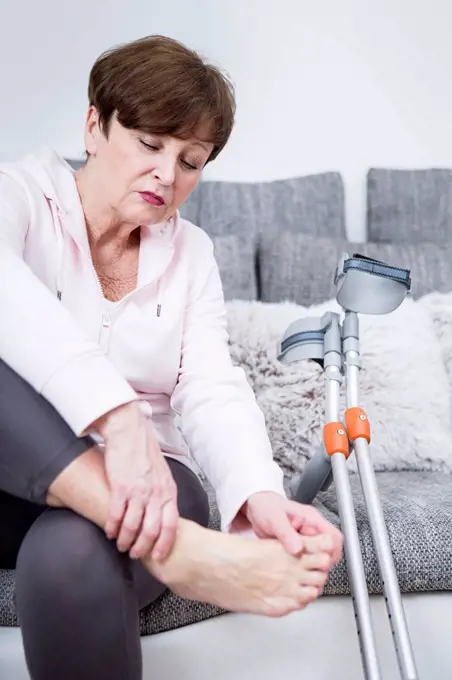 Senior woman with crutches sitting on couch, checking her ankle