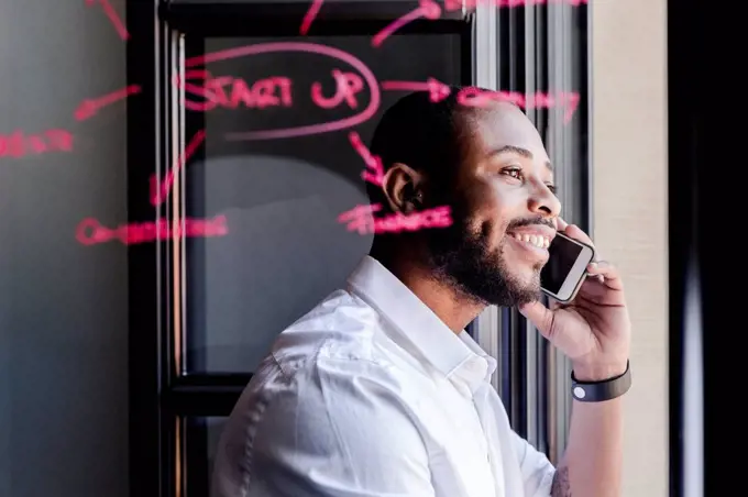 Smiling businessman on cell phone in office with writing on windowpane