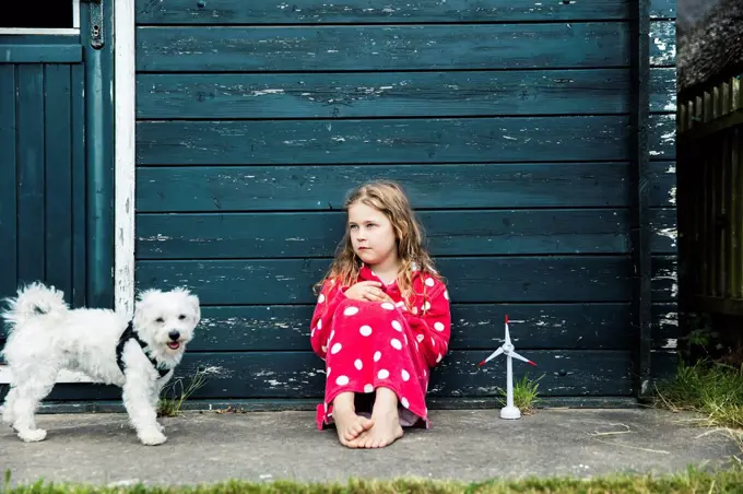 Girl wearing bathrobe outdoors sitting at wooden hut with toy wind turbine and dog