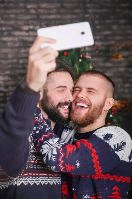Portrait of laughing gay couple taking selfie with smartphone at Christmas time at home