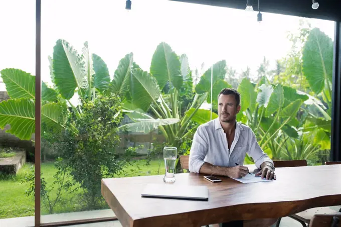 Mature man sitting at table in front of lush garden, writing