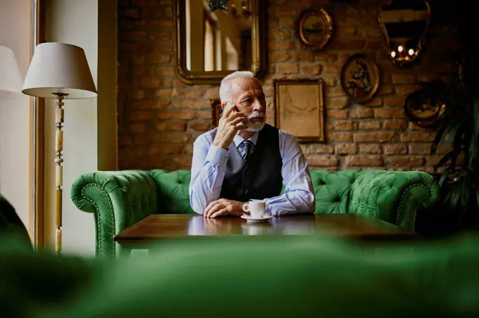 Elegant senior man sitting on couch in a cafe on cell phone