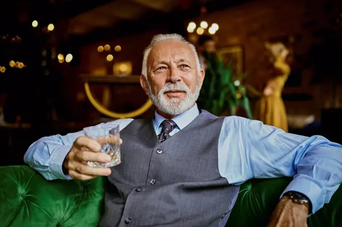 Portrait of elegant senior man sitting on couch in a bar holding tumbler