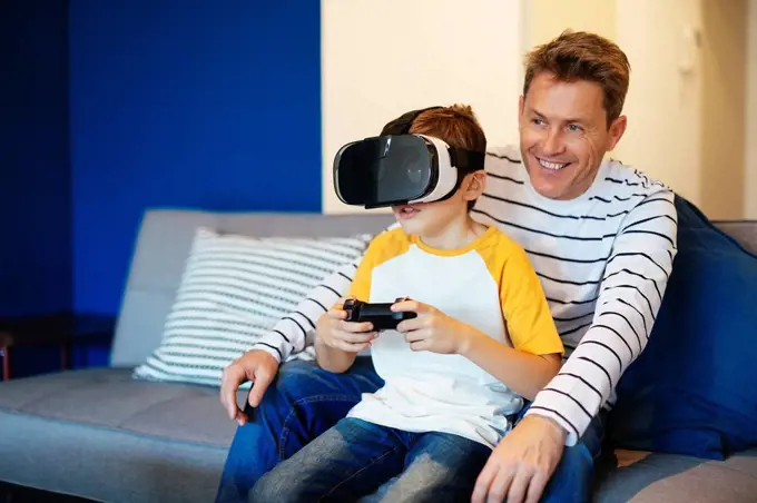 Boy wearing VR glasses playing video game with father on couch at home