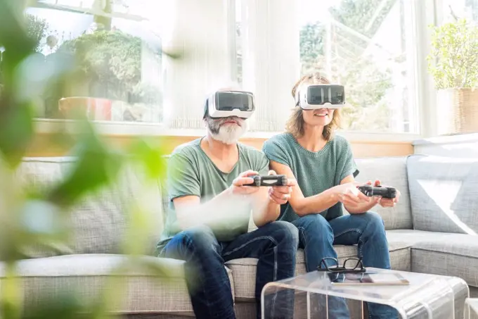 Mature couple sitting on couch at home wearing VR glasses playing video game