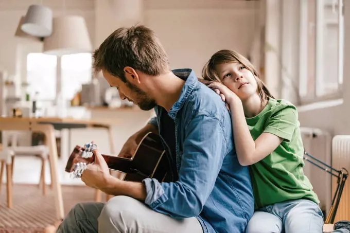 Son leaning against father playing guitar at home