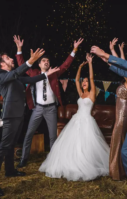 Cheerful wedding couple and friends raising their arms while confetti falling over their heads on a night party outdoors