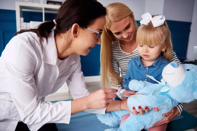 Doctor and girl with mother vaccinating teddy in medical practice
