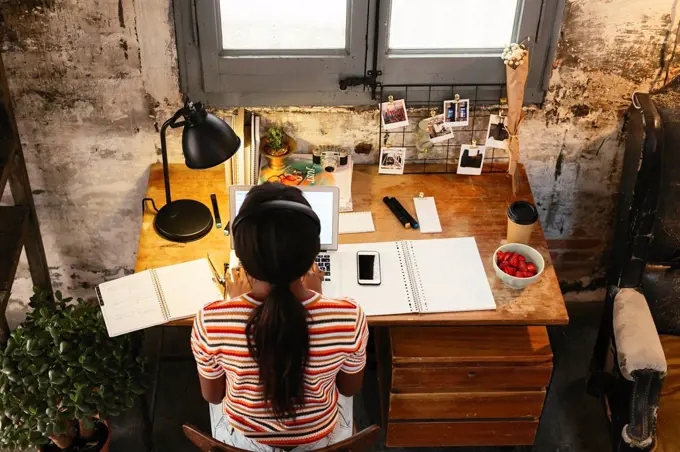 Back view of young woman sitting at desk in a loft working on laptop seen from above