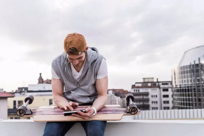 Smiling man sitting in the city with skateboard using cell phone