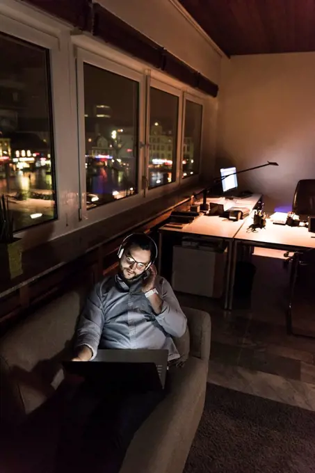 Businessman in office sitting on the couch at night using laptop and headphones