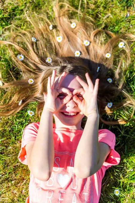 Smiling girl lying on grass in spring with daisies on hair forming spectacles with fingers