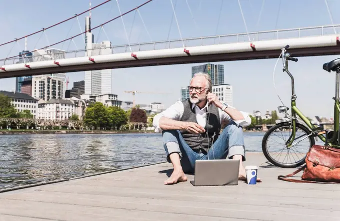 Mature man with laptop, earbuds and bicycle sitting at the riverside in the city