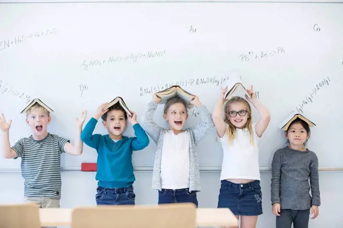 Happy pupils with books above their heads standing at whiteboard with formulas in class