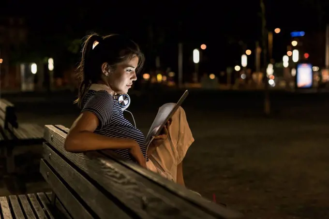 Young woman sitting on bench at night using tablet