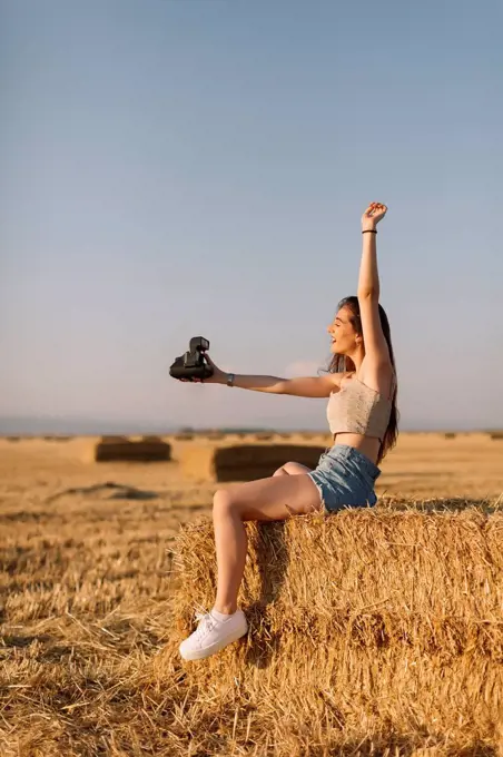 Young woman sitting on straw bale taking selfie with instant camera