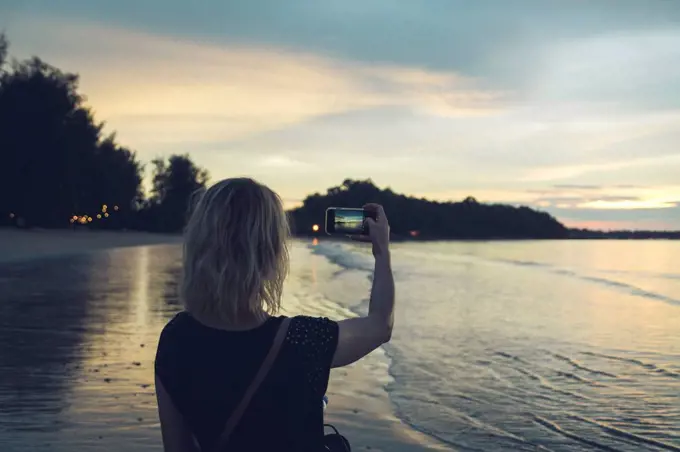Thailand, Khao Lak, back view of woman taking photo with cell phone on the beach at sunset