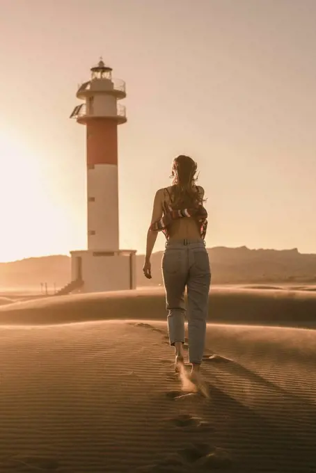 Spain, back view of young woman walking to lighthouse at sunset