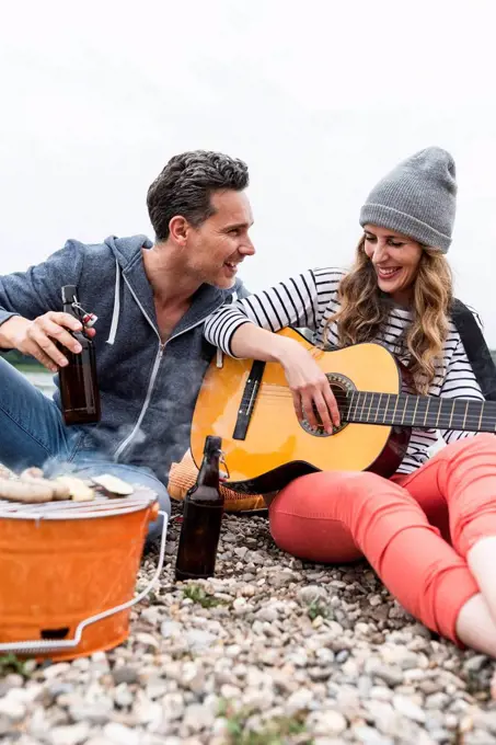 Happy couple with beer bottles, guitar and grill relaxing on pebble beach