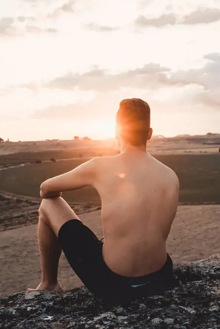 Shirtless young man sitting on rock, watching sunset