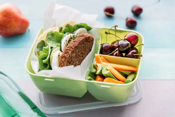 Healthy school food in a lunch box, vegetarian sandwich with cheese, lettuce, cucumber, egg and cress, sliced carrot and celery, cherries and pear