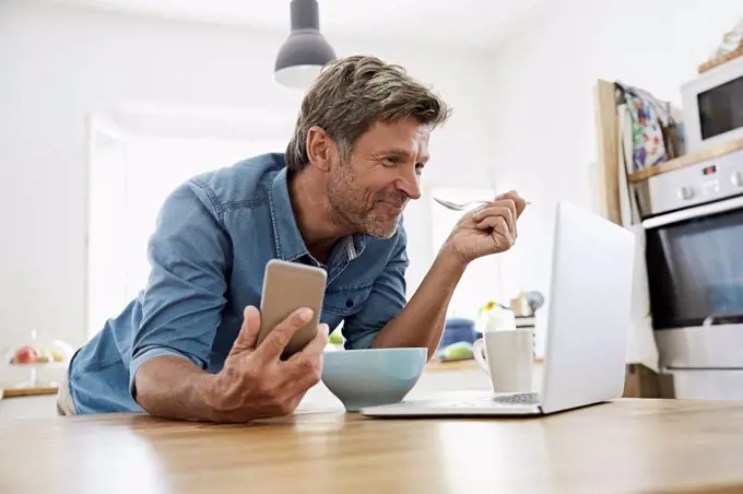Mature man in his kitchen having breakfast, while checking his laptop