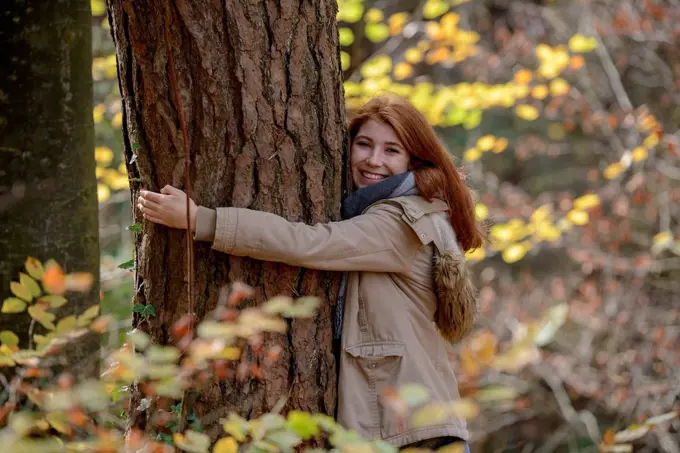 Portrait of happy teenage girl hugging tree trunk in autumnal forest