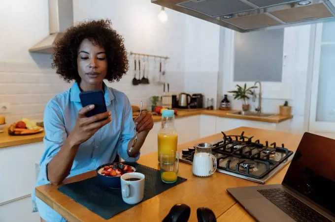 Woman having breakfast in her kitchen, using smartphone