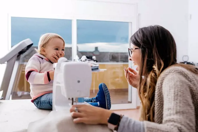 Happy mother with little daughter at home using sewing machine