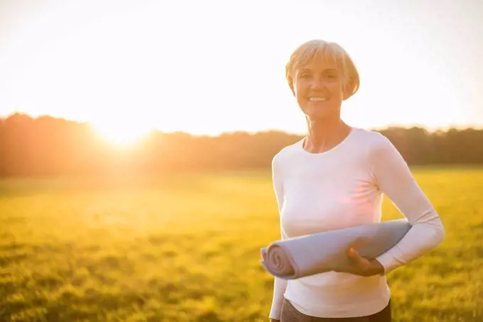 Portrait of smiling senior woman holding yoga mat on rural meadow at sunset