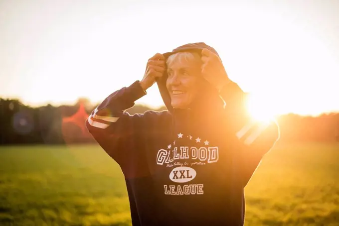 Smiling senior woman wearing a hoodie standing on rural meadow at sunset