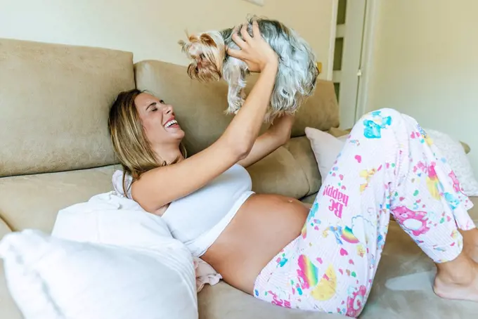 Happy pregnant woman playing with her dog on the couch at home