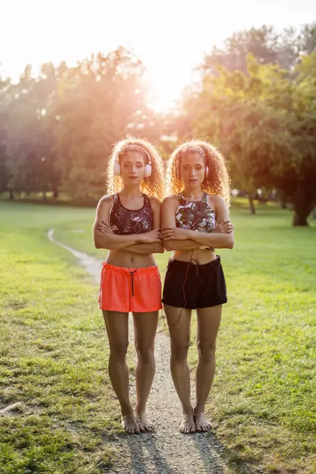 Portrait of twin sisters standing side by side in a park listening music with headphones