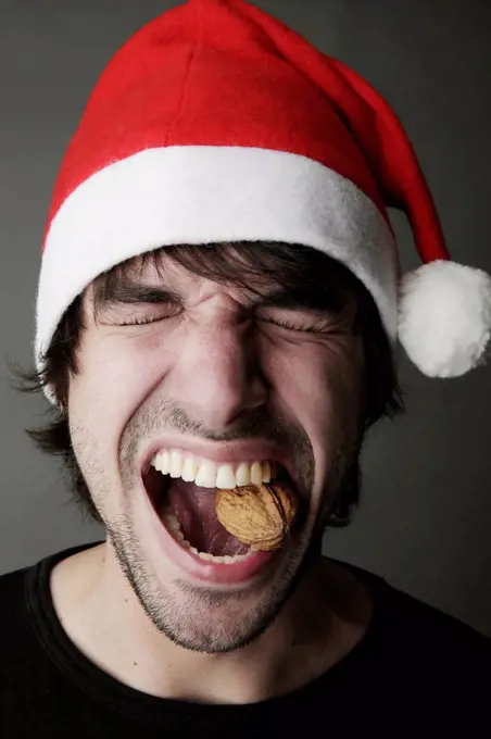 Young man with Santa hat trying to crack a walnut with his teeth, studio shot
