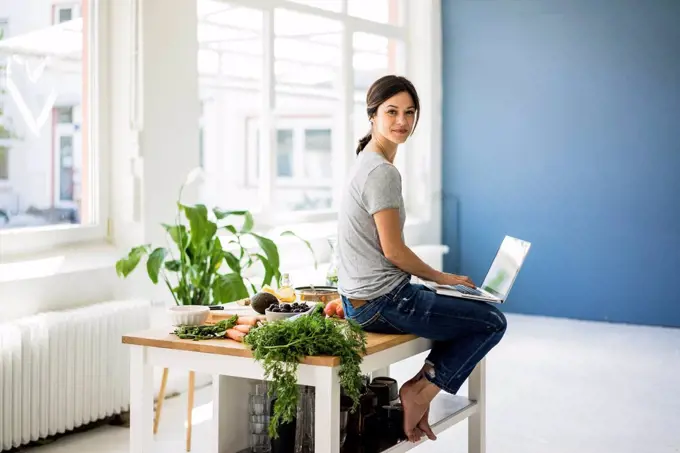 Woman sitting on kitchen table, searching for healthy recipes, using laptop
