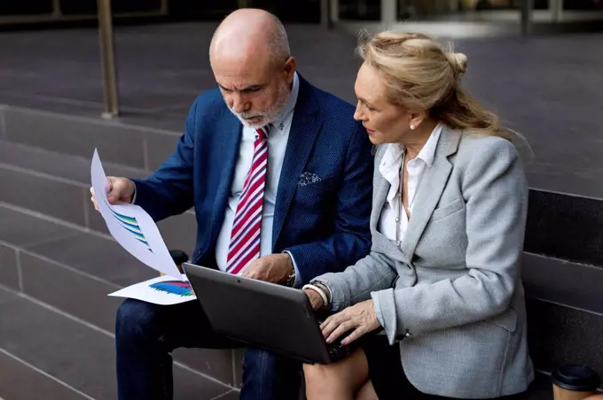 Senior businessman and businesswoman sitting on stairs with laptop and charts