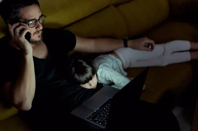 Father talking on cell phone on couch at night with daughter lying next to him