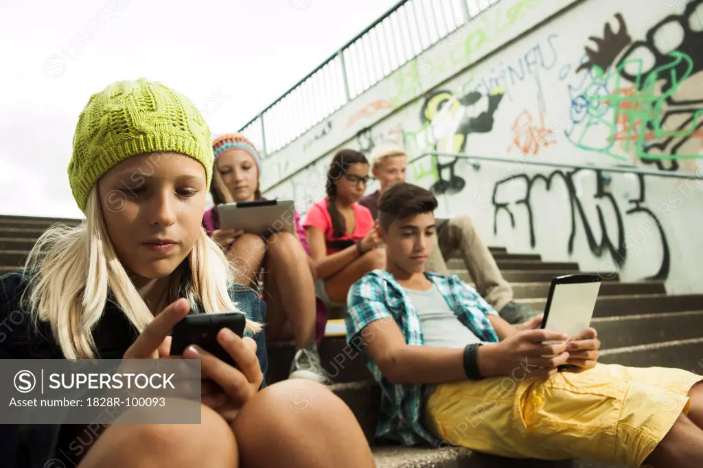 Group of children sitting on stairs outdoors, using tablet computers and smartphones, Germany. 08/31/2013