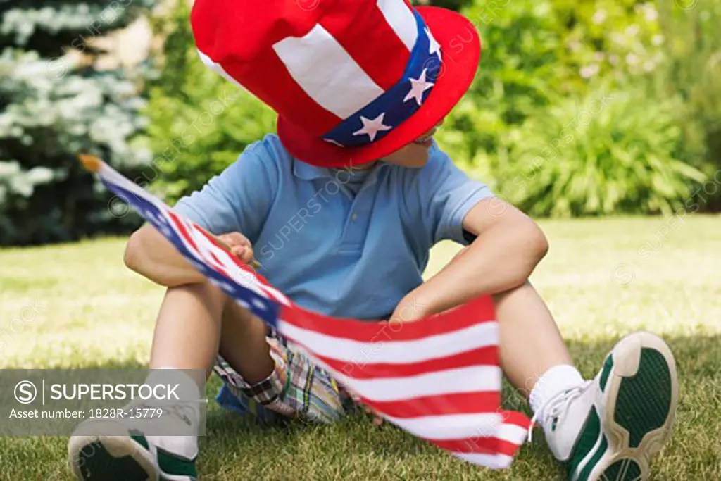 Boy Wearing Large Stars and Stripes Hat, Holding American Flag, Sitting on Grass   