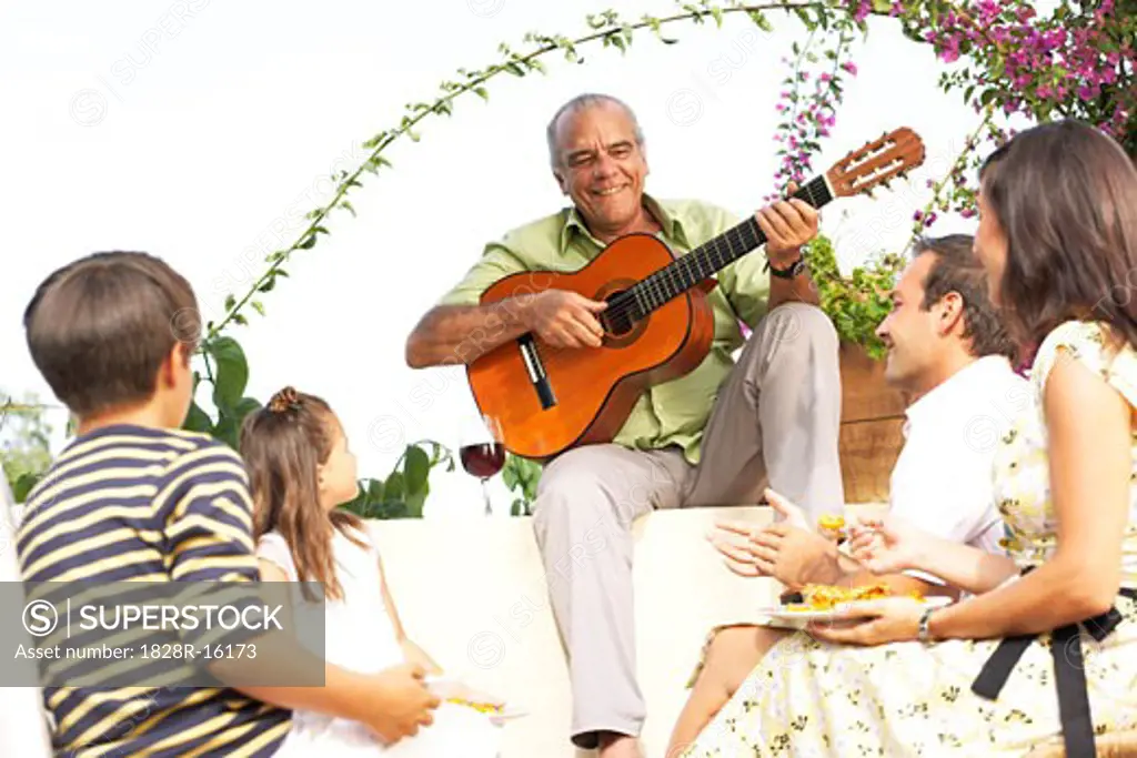 Family Sitting Outdoors Listening to Man Playing Guitar   