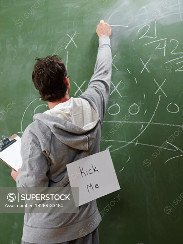 Man at Blackboard with Kick Me Sign on Back   