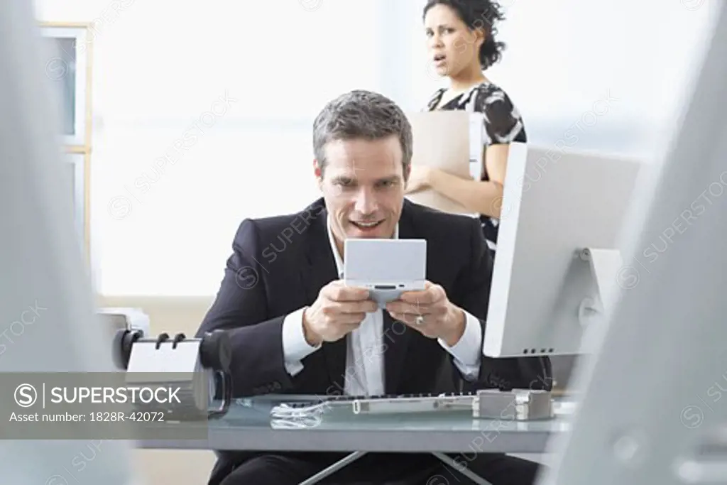 Co-worker looking at Businessman Playing Handheld Video Game at Desk   