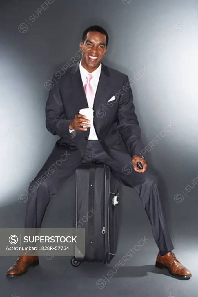 Businessman Sitting on a Suitcase, Holding a Cell Phone and a Cup of Coffee   