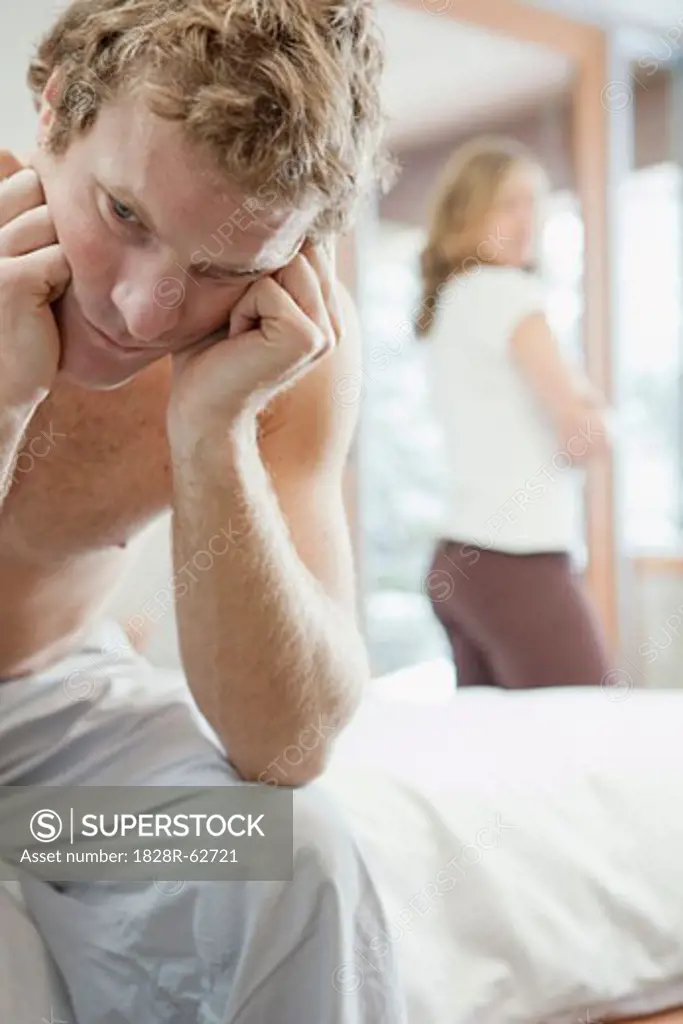 Man Sitting on Edge of Bed and Woman Standing by Window in Background