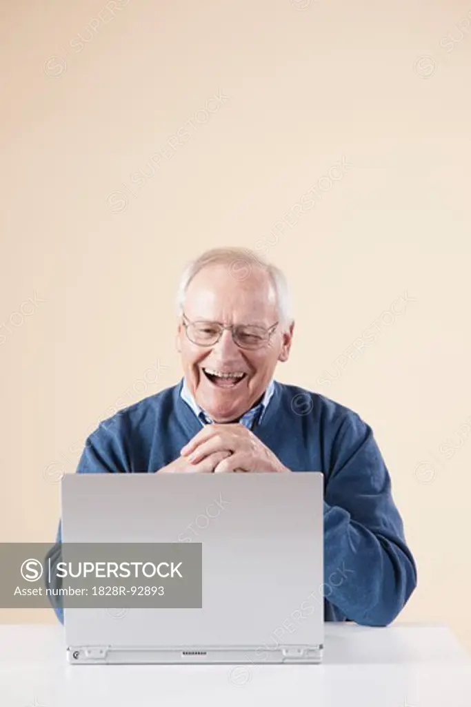 Senior Man Sitting at Table looking at Laptop Computer Laughing, Studio Shot on Beige Background