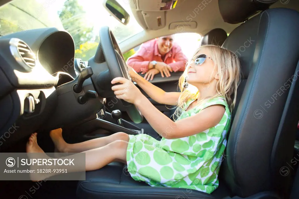 Little girl sitting in Driver's seat of car, pretending to be old enough to drive as her smiling father watches on on a sunny summer evening in Portland, Oregon, USA
